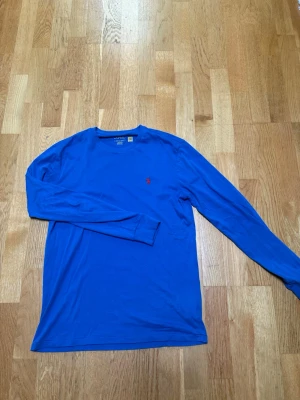 Blå långärmad topp från Polo Ralph Lauren - Snygg blå långärmad topp från Polo Ralph Lauren med klassisk rund halsringning och den ikoniska lilla röda logon broderad på bröstet. Tillverkad i mjuk bomull som känns skön mot huden. Perfekt till jeans eller kjol för en clean look.
