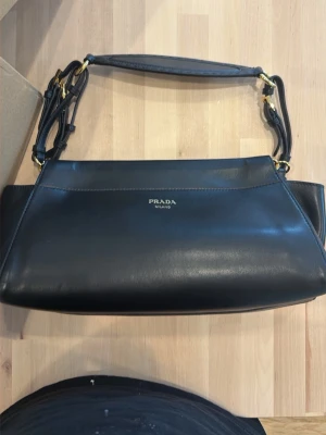 Svart handväska  - Snygg svart handväska  med guldfärgade metalldetaljer och justerbart axelband. Väskan har en stilren, rektangulär form och är tillverkad i slätt läder. Diskret Prada-logo framtill. Perfekt accessoar för en trendig look.