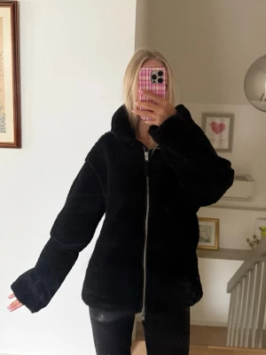 Svart teddyjacka med dragkedja - Mysig svart teddyjacka med stor krage och dragkedja framtill. Jackan har en avslappnad passform och fluffigt material som ger en trendig look. Perfekt för kyliga dagar när du vill vara både varm och snygg. Passar till det mesta i garderoben.