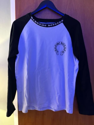 Chrome Hearts långärmad svartvit tröja - Säljer en långärmad svartvit tröja från Chrome Hearts med kontrastärmar och stora logotryck både fram och bak. Tröjan har även tryck på ärmarna och textdetalj i halsen. Perfekt för dig som gillar streetwear och vill sticka ut.
