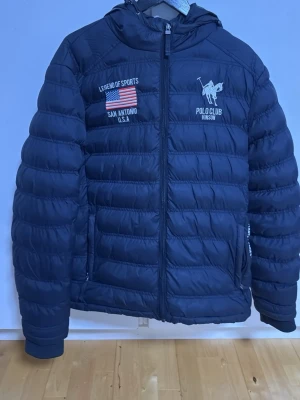 Mörkblå dunjacka med huva - Snygg mörkblå dunjacka med huva och dragkedja framtill. Jackan har broderade detaljer på bröstet, inklusive text och en amerikansk flagga. Quiltad design som ger en sportig look och håller värmen under kalla dagar.
