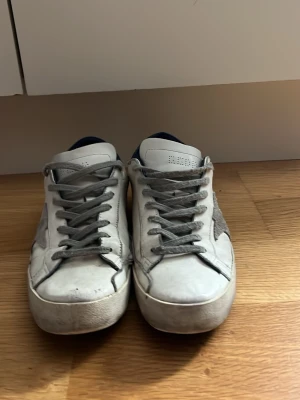 Vita sneakers från Golden Goose - Säljer ett par sneakers från Golden Goose med grå detaljer. Skorna har en klassisk låg siluett och är tillverkade i läder med mockadetaljer på sidan. Perfekta för en avslappnad och trendig stil. Jag säljer för 749kr eftersom jag tycker att det är ett bra pris eftersom kvaliten inte är som ny men det är sjukt enkelt att limma lite så ser man ingen skillnad eller ge den en tvätt kommer göra stor skillnad. De har funkat super för mig men säljer för jag har andra skor. Box finns fråga bara.
