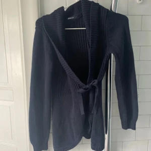 Svart stickad kofta med knyt från Gina Tricot - Mysig svart stickad kofta från Gina Tricot med långa ärmar och bred ribbad krage. Koftan har knytband framtill och är perfekt för lager-på-lager. Snyggt ribbad upptill och slätstickad nedtill för en cool kontrast.