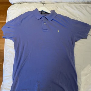 Polo Ralph Lauren - Snygg blå pikétröja från Polo Ralph Lauren med klassisk krage och två knappar framtill. Ikoniska gula loggan broderad på bröstet. Tillverkad i mjuk bomull och har korta ärmar. Perfekt för en clean och stilren look. Hör av dig vid fler frågor