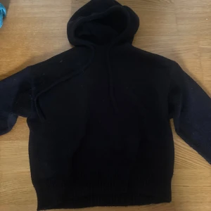 Svart stickad hoodie från Zara - Svart stickad hoodie från Zara i storlek S. Tröjan har huva med snörning och långa ärmar. Perfekt för kyliga dagar och enkel att matcha med det mesta. Ribbstickade muddar vid ärmslut och nederkant ger en snygg finish.
