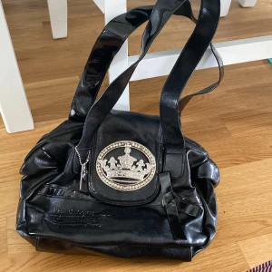 Svart handväska från Friis & Company - Cool svart handväska i glansigt skinnmaterial från Friis & Company. Väskan har en stor silverfärgad krona med stenar på framsidan, dekorativ rosett och dragkedja upptill. Perfekt för dig som gillar statement-detaljer och vill sticka ut.