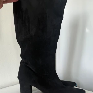 Svarta mockastövlar med klack - Säljer ett par svarta stövlar i fuskmocka med hög klack 8cm och spetsig tå. De når upp under knät. Sula i syntetmaterial. Skaftvidd 40 cm. Strl 38. Mycket fint skick!