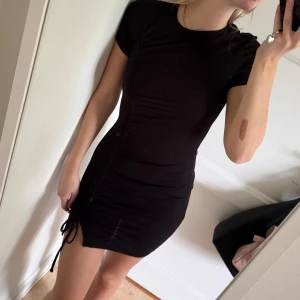 Svart ribbad kortklänning XS från H&M - Supersnygg svart ribbad kortklänning från H&M i storlek XS. Klänningen har korta ärmar, rund hals och cool snörningsdetalj på sidan som ger en extra edge. Materialet är mjukt och stretchigt, perfekt för dig som gillar en tight passform och vill ha en enkel men trendig look.