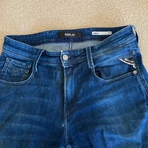 Replay Anbass blå jeans  - Snygga blå jeans från Replay, modell Anbass med slim fit. Klassisk femficksdesign, normal midja och raka ben. Jeansen är tillverkade i mjukt denimtyg och har subtila slitningar för en cool look. Perfekta för dig som gillar stilrena och moderna jeans.