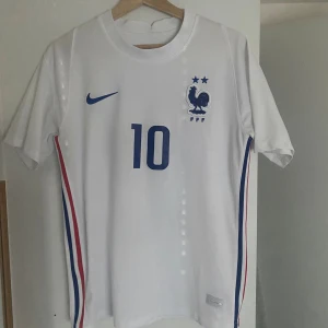 Frankrike Mbappé 10 Nike fotbollströja - Säljer en officiell Frankrike fotbollströja från Nike med nummer 10 och Mbappé på ryggen. Tröjan är vit med blå detaljer, broderat FFF-emblem och två stjärnor på bröstet. Sidorna har rödblå ränder. Tillverkad i lätt och ventilerande material, perfekt för match eller träning. Kom dm för fler frågor eller bilder. Kan gå ner pris vid snabb affär. 