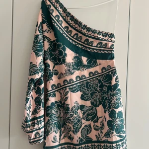 Blommig oneshoulder klänning - Snygg kortklänning med blommigt mönster i mörkgrönt och ljusrosa. Klänningen har en ärm och en axel bar, vilket ger en trendig oneshoulder-look. Perfekt för dig som vill sticka ut med en unik och cool stil.