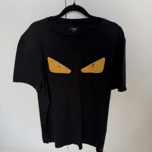 Svart Fendi t-shirt med gula ögon - Svart t-shirt från Fendi med ikoniska gula ögon framtill. Klassisk rund halsringning och korta ärmar. Tillverkad i mjuk bomull för en skön känsla. Diskret Fendi-detalj nertill på sidan. Perfekt för dig som vill sticka ut med en cool designerlook.