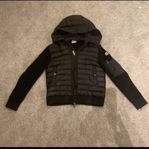 Moncler cardigan  - Säljer en svart cardigan från Moncler med stickade ärmar och huva. Jackan har dragkedja framtill, två fickor med knappar och en extra ficka på ärmen. Klassisk Moncler-logga på ena ärmen. Perfekt för kyliga dagar och riktigt snygg streetstil.