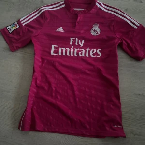 Rosa Real Madrid fotbollströja Adidas - Säljer en officiell Real Madrid fotbollströja från Adidas i en snygg rosa färg med vita detaljer och ränder på axlarna. Tröjan har klubbmärke, Fly Emirates-tryck och La Liga-märke på ärmen. Tillverkad i lätt och ventilerande climacool-material.