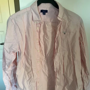 Ljusrosa skjorta från Gant - Säljer en stilren ljusrosa skjorta från Gant med klassisk krage och knäppning framtill. Skjortan har lång ärm och diskret Gant-logga broderad på bröstet. Perfekt för dig som gillar en clean och fräsch look.