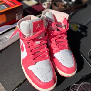 Nike Air Jordan 1 rosa - Säljer ett par Nike Air Jordan 1 i en riktigt snygg färgkombo av rosa och vitt. Skorna har klassisk high-top design, perforerad tå och ikoniska swoosh-loggan på sidan. Tillverkade i skinn med rosa snörning och vit sula. 