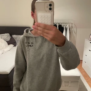Grå hoodie från Gina Tricot med tryck - Grå hoodie från Gina Tricot med svart texttryck 'JE M'EN AMOUR' och en stjärna på bröstet. Tröjan har huva med snörning och är långärmad. Mjuk insida och avslappnad passform, perfekt för chill dagar.
