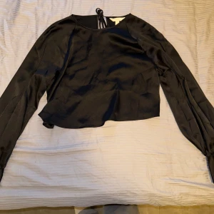 Svart satinblus från H&M med knyt - Säljer en svart blus från H&M i glansigt satinmaterial. Blusen har långa puffiga ärmar med knappar vid ärmslut och en snygg knytning i nacken. Croppad modell. 