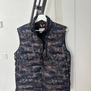 Camo dunväst från SWIMS - Snygg dunväst från SWIMS i camouflage-mönster med dragkedja framtill och hög krage. Västen har flera fickor och är perfekt för lager-på-lager. Färgerna går i mörkblått, grått och brunt. Perfekt för dig som vill ha en cool och funktionell look. Fits medium 