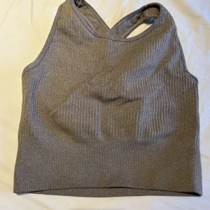 Grå ribbad linnetopp med brottarrygg - Säljer en grå ribbad linnetopp med brottarrygg. Toppen har en sportig look med smala axelband och ärmlös design, perfekt för träning eller chill dagar. Materialet är stretchigt och följsamt för en bekväm passform.