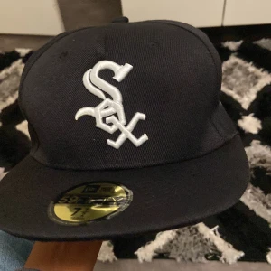 Svart New Era White Sox keps 59FIFTY - Snygg svart keps från New Era, modell 59FIFTY, med broderad vit White Sox-logga framtill och MLB-logga baktill. Klassisk rak skärm och passform som sitter perfekt. Kepsen är tillverkad i polyester och har märkets detaljer invändigt.