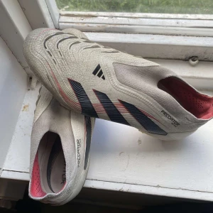 Adidas Predator fotbollsskor beige/svart - Säljer ett par Adidas Predator fotbollsskor i beige med svarta och röda detaljer. Skorna har en strumpliknande passform utan snörning, mönstrad ovandel och dobbar för gräsplan. Snygg design med klassiska Adidas-ränder och Predator-tryck på sulan. Tecken på användning, användts sparsamt! Pris ej hugget i sten, snabbaffär osv. 41 1/3. Lägg bud på skorna. Pris diskuteras!