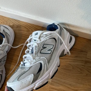 New Balance vita sneakers mesh silver - Säljer ett par klassiska New Balance sneakers i vitt med silver- och marinblå detaljer. Skorna har meshpaneler för extra andningsförmåga, snörning och en robust sula i svart och vitt. Perfekta för dig som gillar sportig och clean stil. Pris kan diskuteras 