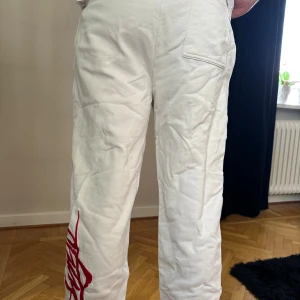 Vita mjukisbyxor med röd logga - Vita mjukisbyxor från Hugo med röd och svart logotyp på höger lår och stor röd broderad text längs vänster ben. Byxorna har snörning i midjan, resår vid benslut och är tillverkade i mjuk bomull. Perfekta för en chill och avslappnad stil. Pris kan diskuteras 