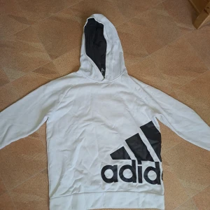 Vit hoodie från Adidas med svart logga - Säljer en vit hoodie från Adidas med stor svart logga och ikoniska ränder på framsidan. Tröjan har huva med svart insida och långa ärmar. Perfekt för dig som gillar sportig och avslappnad stil.