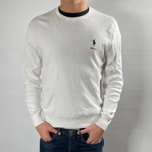 Vit tröja från Polo Ralph Lauren - Vit långärmad tröja från Polo Ralph Lauren med klassisk rund halsringning. Tröjan har broderad logga i marinblått och rött på bröstet. Tillverkad i mjuk bomull, perfekt för en clean och stilren look.