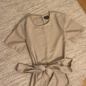 Beige blus från Bubbleroom - Stilren beige blus från Bubbleroom med rund halsringning, korta ärmar och ett brett knytband i midjan. Blusen har en enkel knapp i nacken och är tillverkad i ett mjukt, lätt material som ger en snygg passform.