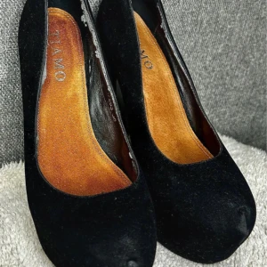 Svarta mocka pumps från Tiamo - Snygga svarta pumps från Tiamo i mocka med hög klack och plattform. Skorna har rund tå och en klassisk siluett som ger extra höjd. Insidan är i brunt material och utsidan är helt i svart mocka. Perfekta för dig som vill sticka ut lite extra. Ett par eleganta svarta pumps från Tiamo i storlek 36. Klassiska pumps i mockaimitation, knappt använda. slitage beror på att dom legat i en låda tillsammans med andra skor samt inte använts.