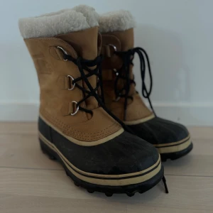 Sorel vinterboots med foder - Bruna och svarta vinterboots från Sorel med vitt, fluffigt foder upptill. Skorna har snörning framtill, grov sula och är tillverkade i skinn och gummi. Perfekta för kalla dagar när du vill hålla fötterna varma och torra.