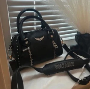 Zadig & Voltaire väska - Cool svart handväska från Zadig & Voltaire med silvriga detaljer, nitar och logga framtill. Väskan har både handtag och axelrem med texten VOLTAIRE. Perfekt för dig som vill ha en edgy och snygg accessoar.