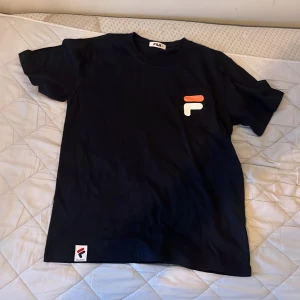 Svart t-shirt från FILA med logga - Svart t-shirt från FILA med klassisk logga i rött, vitt och marinblått på bröstfickan och en liten etikett nertill. Tillverkad i 100% bomull och har en enkel, stilren design med korta ärmar och rund hals.