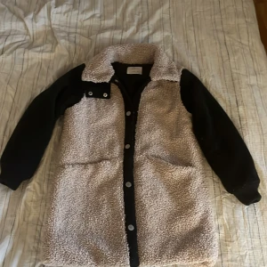  teddyjacka från Zara - Säljer en lång teddyjacka från Zara i beige med svarta ärmar och detaljer. Jackan har stora fickor, knappar framtill och en mysig krage. Perfekt för kyliga dagar och riktigt snygg till streetwear-stil.