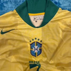 Brasilien Vini Jr. 7 Nike fotbollströja - Gul Brasilien fotbollströja från Nike med gröna detaljer, nummer 7 och VINI JR. på ryggen. Broderat CBF-märke på bröstet och Nike-logga. Tillverkad i Dri-Fit material som andas, perfekt för match eller träning.