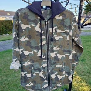 Camouflage parkajacka från H&M Divided - Snygg parkajacka i camouflage från H&M Divided, storlek 36. Jackan har en svart huva, dragkedja framtill och flera fickor. Materialet är bomull och polyester, perfekt för dig som gillar streetstyle och vill sticka ut med mönster. Passformen är normal och jackan är långärmad.