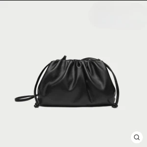 Svart axelväska med snörning - Stilren svart axelväska i mjukt skinnimitation med cool snörningsdetalj upptill. Väskan har en minimalistisk design med släta ytor och tunna axelband, perfekt för dig som gillar en clean och modern look.