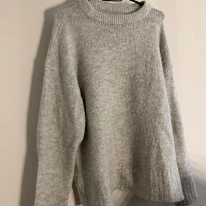 Ljusgrå stickad tröja - Mysig ljusgrå stickad tröja med rund hals och avslappnad passform. Perfekt för kyliga dagar och enkel att matcha med jeans eller kjol. Tröjan har långa ärmar och ribbade muddar vid hals, ärmslut och nederkant.