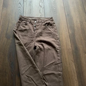 Bruna wide jeans från Never Denim - Bruna jeans från Never Denim med hög midja och straight fit. Klassisk femficksmodell med knäppning framtill och läderpatch bak i midjan. Aldrig använda.