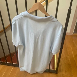 Ljusblå Lacoste slim fit piké - Fräsch ljusblå pikétröja från Lacoste i slim fit-modell. Klassisk krage, korta ärmar och knäppning framtill. Tröjan är gjord i mjuk bomull och har den ikoniska gröna krokodilloggan på bröstet. Perfekt för dig som gillar stilren och enkel design.