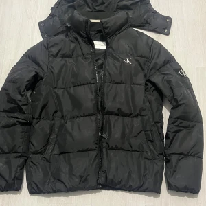 Calvin Klein Jacka - Svart pufferjacka från Calvin Klein Jeans med avtagbar huva och dragkedja framtill. Jackan har diskret CK-logga på bröstet och ärmen, samt två sidofickor. Perfekt för kalla dagar och har en clean, stilren look. Lite smutsig men en tvätt så är den stilren annars inga defekter. Nypris runt 3000kr