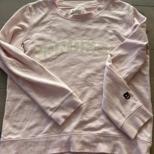 Ljusrosa sweatshirt från Bondelid XS - Superskön ljusrosa sweatshirt från Bondelid i bomull. Klassisk modell med rund hals, ribbade muddar och tryckt logga framtill. Liten patch på ena ärmen ger extra detalj. Perfekt för en chill och avslappnad stil.
