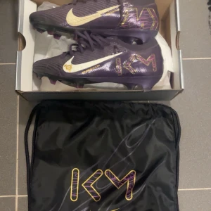 Nike Air Zoom Mercurial fotbollsskor lila - Säljer ett par Nike Air Zoom Mercurial fotbollsskor i mörklila med guldiga och vita detaljer. Skorna har snörning, mönstrad sula för bra grepp och coola KM-loggor på sidorna. Kommer med tillhörande svart dustbag med matchande tryck. Perfekta för dig som vill sticka ut på planen! Använda 2-3 gånger