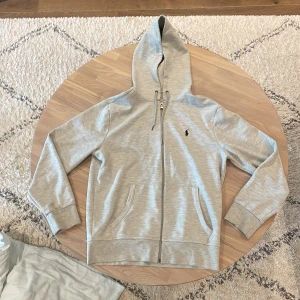 Grå hoodie från Polo Ralph Lauren - Snygg ljusgrå hoodie från Polo Ralph Lauren med dragkedja och klassisk logga på bröstet. Tröjan har huva med snörning, två fickor framtill och ribbade muddar. Perfekt för chill dagar eller när du vill ha en clean look. Storlek S passar Xs