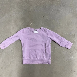 Ljuslila sweatshirt från Lindex Kids - En enkel och mjuk ljuslila sweatshirt från Lindex Kids i storlek 110. Tröjan har rund hals, ribbade muddar vid ärmslut och nederkant samt är tillverkad i bomull. Perfekt för en chill och avslappnad stil.