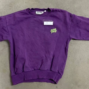 Lila sweatshirt från Junkyard - Cool lila sweatshirt från Junkyard med limegrönt tryck på bröstet. Tröjan har rund hals, ribbade muddar och är tillverkad i mjuk bomull. Perfekt för dig som gillar streetwear och vill sticka ut med färg.
