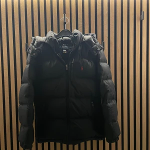 Svart pufferjacka från Polo Ralph Lauren - Svin snygg svart pufferjacka från Polo Ralph Lauren med huva, dragkedjor framtill och röd broderad logga på bröstet. Jackan har quiltad design och elastiska muddar vid ärmslut. Perfekt för kalla dagar när du vill hålla stilen. Inga hål eller liknande, köpt för 3395kr, barn storlek, L (14-16) (160-171), men passar även xs skulle jag säga! Skicka om ni vill ha mått så löser jag! hör av er om ni har någon fundering!😊
