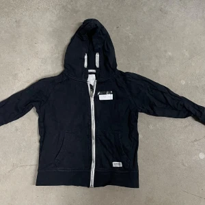 Svart hoodie med dragkedja från 157 - Svart hoodie från 157 Denim Makers med vit dragkedja och grå snören i huvan. Tröjan har känguruficka framtill och är långärmad. Perfekt för chill dagar och enkel att matcha med jeans eller joggers.